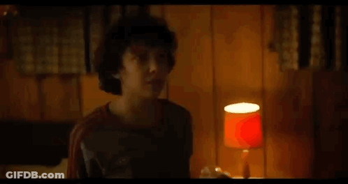 A Young Boy Is Standing In A Room With A Lamp And A Wooden Wall . GIF