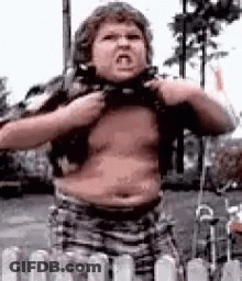 A Young Boy Is Standing In Front Of A Fence With His Shirt Off And Making A Funny Face . GIF