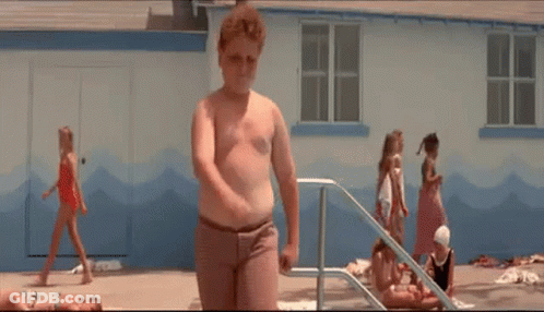 A Young Boy Is Standing In Front Of A Swimming Pool While A Group Of Children Are Playing In The Water . GIF