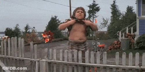 A Young Boy Is Standing In Front Of A Wooden Fence And Looking Over It . GIF