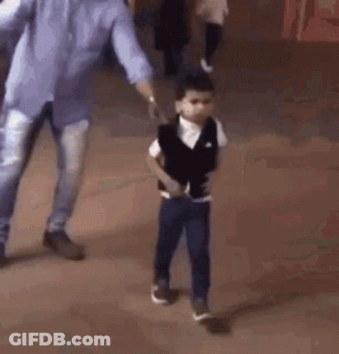 A Young Boy Is Standing On A Tiled Floor With His Hands In His Pockets And A Man Walking Behind Him . GIF