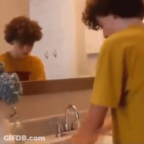 A Young Boy Is Washing His Hands In A Bathroom Sink While Looking At Himself In The Mirror . GIF