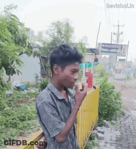 A Young Boy Smoking A Cigarette In Front Of A Yellow Fence With The Website Bavishni.in Written On The Bottom GIF