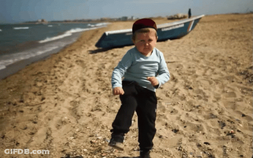 A Young Boy Stands On A Beach With A Boat In The Background GIF