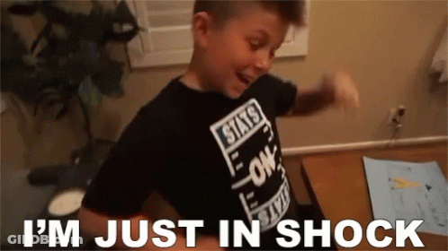 A Young Boy Wearing A Black Shirt That Says I 'M Just In Shock Is Standing In Front Of A Desk . GIF