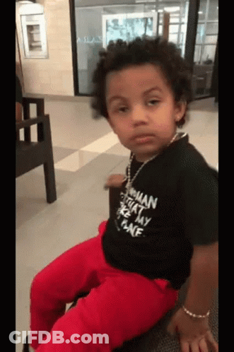 A Young Boy Wearing A Black Shirt That Says ' Woman ' On It Is Sitting On A Chair . GIF