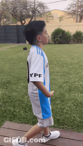 A Young Boy Wearing A Blue And White Ypf Jersey Walks Across A Wooden Deck GIF
