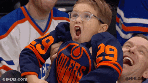 A Young Boy Wearing A Blue Oilers Jersey Is Being Held By A Man GIF