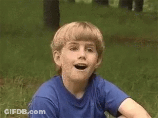 A Young Boy Wearing A Blue Shirt Is Sitting In The Grass With His Mouth Open GIF