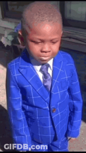 A Young Boy Wearing A Blue Suit And Tie Is Standing In Front Of A Window . GIF