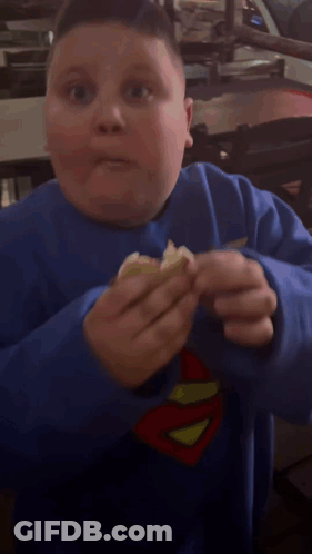 A Young Boy Wearing A Blue Superman Sweatshirt Is Eating Something GIF