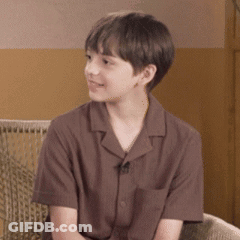 A Young Boy Wearing A Brown Shirt Is Sitting On A Chair . GIF
