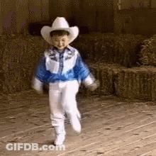 A Young Boy Wearing A Cowboy Hat And A Blue Jacket Is Walking On A Wooden Floor . GIF