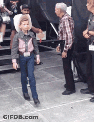 A Young Boy Wearing A Cowboy Hat And Suspenders Is Walking On The Sidewalk GIF