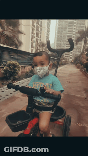 A Young Boy Wearing A Face Mask Is Riding A Tricycle GIF