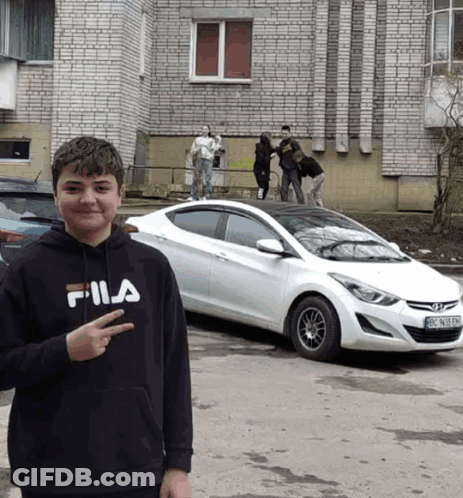 A Young Boy Wearing A Fila Sweatshirt Stands In Front Of A White Car GIF