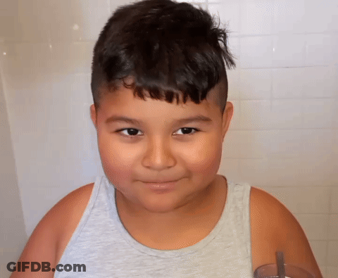 A Young Boy Wearing A Grey Tank Top Is Smiling And Holding A Glass GIF