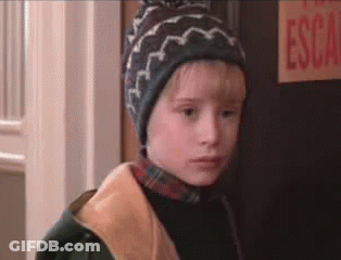 A Young Boy Wearing A Hat And A Jacket Is Standing In Front Of A Door . GIF