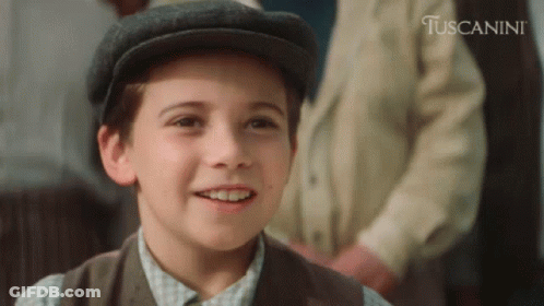 A Young Boy Wearing A Hat Is Smiling In Front Of A Sign That Says Tuscanini GIF