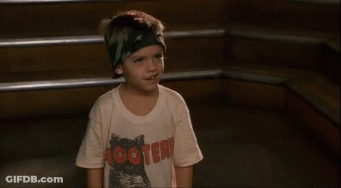 A Young Boy Wearing A Hooters T-Shirt And A Headband Is Standing In Front Of Some Stairs . GIF