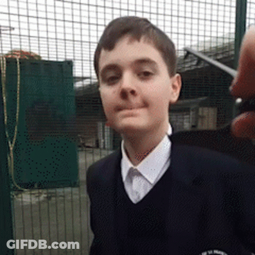 A Young Boy Wearing A School Uniform Stands In Front Of A Fence GIF