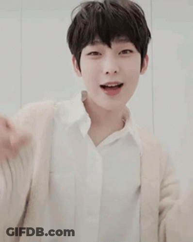 A Young Boy Wearing A White Shirt And A Beige Cardigan Is Smiling And Pointing . GIF