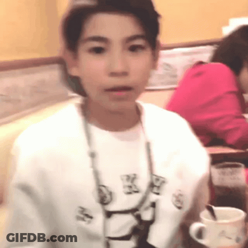 A Young Boy Wearing A White Shirt With A Necklace Around His Neck Is Sitting At A Table In A Restaurant . GIF