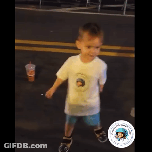 A Young Boy Wearing A White Shirt With A Picture Of A Man On It Is Walking Down A Street GIF