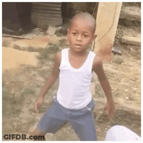 A Young Boy Wearing A White Tank Top And Blue Shorts Is Standing In A Dirt Field . GIF