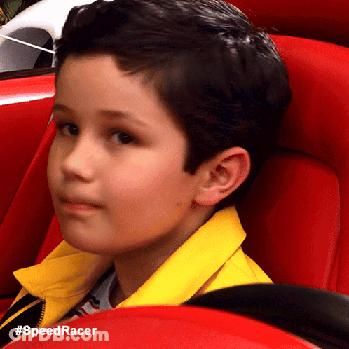 A Young Boy Wearing A Yellow Jacket Is Sitting In A Red Car With The Hashtag #Speedracer GIF
