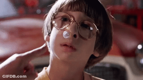 A Young Boy Wearing Glasses Is Blowing A Bubble In His Nose In Front Of A Red Car . GIF