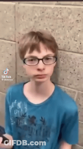 A Young Boy Wearing Glasses Is Standing In Front Of A Brick Wall . GIF