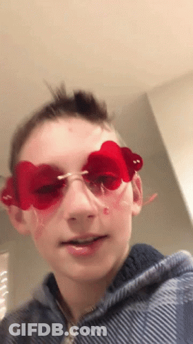A Young Boy Wearing Red Heart Shaped Sunglasses Looks At The Camera . GIF