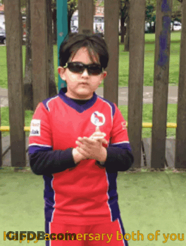 A Young Boy Wearing Sunglasses Holds A Lollipop In Front Of A Fence With The Words Happy Anniversary Both Of You Written GIF