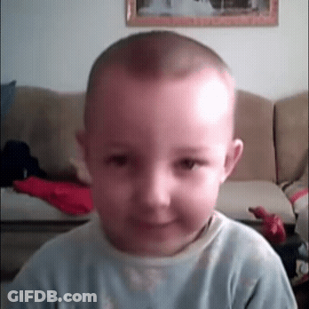 A Young Boy With A Shaved Head Is Smiling In Front Of A Couch GIF
