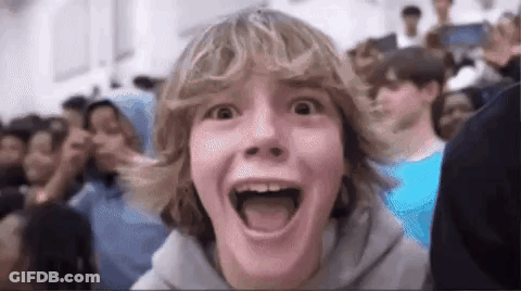 A Young Boy With Blonde Hair Is Making A Funny Face In Front Of A Crowd Of People . GIF