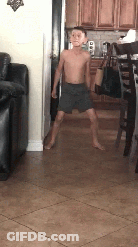 A Young Boy Without A Shirt Is Standing In A Living Room . GIF
