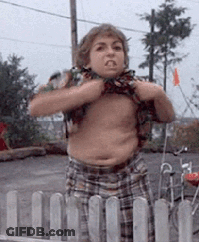 A Young Boy Without A Shirt Is Standing In Front Of A Fence And Making A Funny Face . GIF