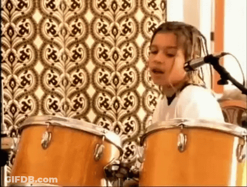 A Young Girl Is Playing Drums And Singing Into A Microphone In Front Of A Patterned Wall . GIF