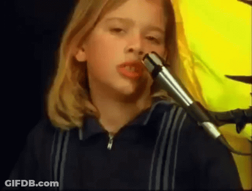 A Young Girl Is Singing Into A Microphone With A Yellow Background Behind Her . GIF