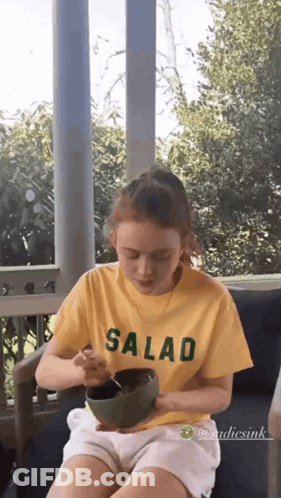 A Young Girl Is Sitting On A Porch Eating A Salad From A Bowl . GIF
