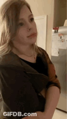 A Young Girl Is Standing In Front Of A Refrigerator And Looking At The Camera . GIF