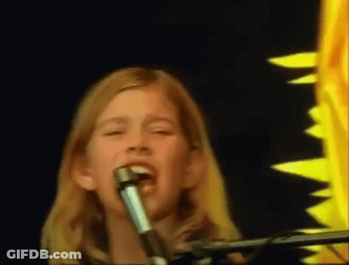 A Young Girl Singing Into A Microphone In Front Of A Sunflower GIF