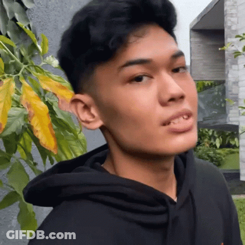 A Young Man In A Black Hoodie Is Standing In Front Of A Tree And Looking At The Camera . GIF