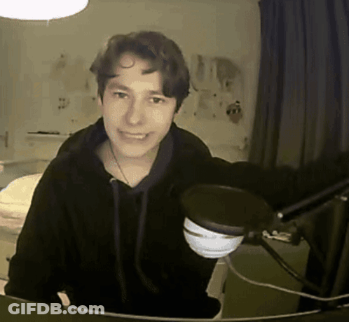 A Young Man In A Black Hoodie Stands In Front Of A Microphone And Smiles GIF