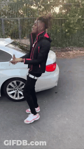 A Young Man In A Black Jacket And Pink Shoes Is Standing In Front Of A White Car GIF