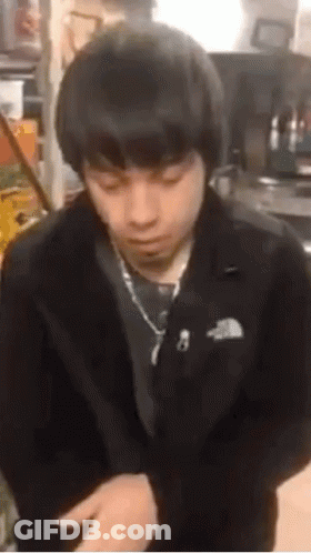 A Young Man In A Black Jacket And Tie Is Sitting In A Kitchen . GIF