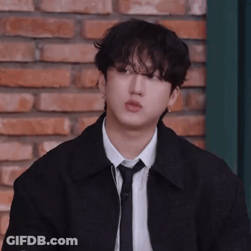 A Young Man In A Black Jacket And Tie Is Standing In Front Of A Brick Wall . GIF