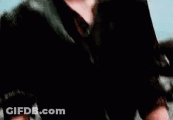 A Young Man In A Black Jacket Is Standing In Front Of A Blue Background . GIF