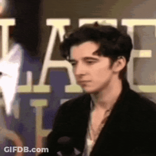 A Young Man In A Black Jacket Is Standing In Front Of A Sign That Says ' A ' On It . GIF